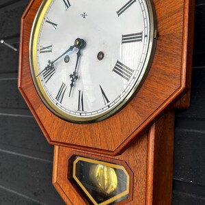 Puede incluir: Un reloj de pared de madera vintage con esfera blanca y números romanos negros. El reloj tiene un bisel dorado y un péndulo visible a través de una ventana de cristal. El marco de madera es de color marrón cálido.