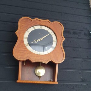 May include: A vintage wooden wall clock with a dark face and gold-colored hands and Roman numerals. The clock has a decorative, scalloped frame and a pendulum in a small, open compartment below. The clock is mounted on a dark, textured wall.