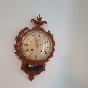 Puede incluir: Un reloj de pared de madera con esfera crema, números romanos y manecillas doradas. El reloj está rodeado por un marco de madera tallada con un diseño de hojas de laurel. Tiene una parte superior decorativa y un área de péndulo en la parte inferior.