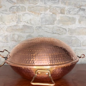 Puede incluir: Una olla de cocina de metal martillado de color cobre con una tapa abovedada. La olla tiene dos asas a los lados y un asa en la parte delantera. La olla está sobre una superficie de madera.