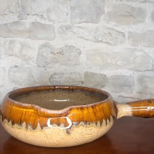 May include: A ceramic soup bowl with a handle, featuring a speckled tan base and a glossy, brown-glazed rim. The bowl has a pouring spout and is set against a textured, light-colored stone wall.
