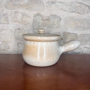 May include: A speckled, off-white ceramic soup pot with a lid and a handle. The pot has a rounded body and a small, round lid with a knob. The handle is curved and extends from the side of the pot. The pot is on a wooden surface.
