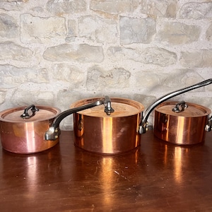 May include: Three copper saucepans with lids and black handles are displayed on a wooden surface. The pans have a polished, reflective finish. The background features a light-coloured stone wall.