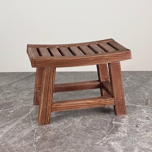 Puede incluir: Un taburete pequeño de madera con acabado marrón oscuro y asiento curvo de listones. El taburete cuenta con cuatro patas robustas conectadas por barras de soporte horizontales, mostrando una textura de veta de madera visible.