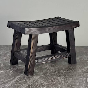 Peut inclure: Un petit tabouret en bois avec une finition carbonisée foncée et une assise incurvée à lattes. Le tabouret présente quatre pieds robustes reliés par des barres de support horizontales, mettant en valeur la texture du grain du bois naturel.