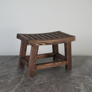 Puede incluir: Un taburete pequeño de madera con un acabado teñido en marrón oscuro y un patrón de veta de madera visible. El asiento presenta un diseño curvo de listones apoyado por cuatro patas robustas conectadas por travesaños horizontales en la base.