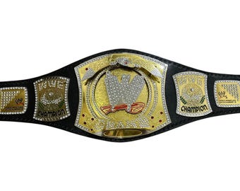 May include: A black leather championship belt featuring a large central gold plate with a silver WWE logo, surrounded by smaller gold side plates. The plates are embellished with clear crystals and red accents, with the text WWE CHAMPION visible.