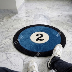 May include: Circular rug designed as a pool ball, with a blue central area, a white circle, and the number 2 in black. It has a black border. The image includes a person's legs and feet wearing white trainers.