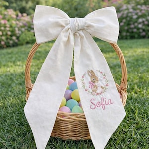 Puede incluir: Una cesta de Pascua de mimbre tejido llena de huevos coloridos, adornada con un gran lazo color crema. La cola del lazo presenta un diseño de conejito bordado dentro de una corona floral y el nombre "Sofia" en escritura rosa.