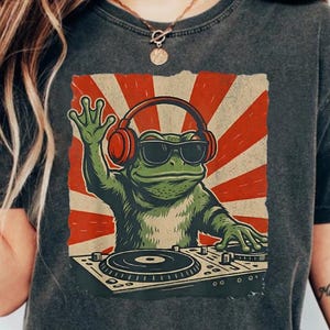 Puede incluir: Camiseta gris oscuro con un DJ rana verde con gafas de sol y auriculares, sobre un fondo de rayos rojos y blancos. La rana está detrás de una mesa de mezclas, saludando. La camiseta es de estilo casual y unisex.