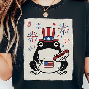 Puede incluir: Camiseta gris oscuro con un dibujo de una rana con un sombrero patriótico y un perrito caliente. La rana tiene una bandera americana en la barriga, con fuegos artificiales en el fondo. La camiseta tiene un aspecto vintage.