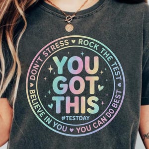 Pode incluir: Camiseta cinza escuro com um design circular que diz "YOU GOT THIS" em cores pastel. O design inclui as frases "Don't Stress, Rock the Test, Believe in You, You Can Do Best" e a hashtag #TestDay.
