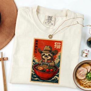 May include: Cream-colored t-shirt featuring a graphic of a sloth wearing a cowboy hat and eating ramen from a red bowl. The graphic has the word "RAMEN" and Japanese characters. A gold necklace and a bowl of ramen are also visible.