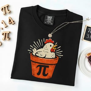 Puede incluir: Sudadera negra con un gráfico de un pollo en una maceta con el símbolo pi. El diseño es en naranja, blanco y rojo. También se ven un collar plateado y una porción de tarta.