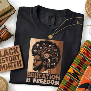 May include: Black t-shirt featuring a graphic of a Black man with a tree-like brain, the text "EDUCATION IS FREEDOM", and a gold necklace. A wooden sign reads "BLACK HISTORY MONTH". Books and a small drum are also visible.