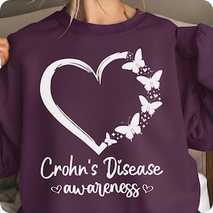 May include: A dark purple sweatshirt with a white heart and butterfly design. The words "Crohn's Disease awareness" are printed in white script below the heart. The design includes small hearts.