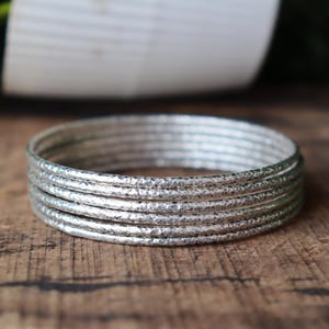 May include: A stack of silver bangles with a hammered texture. The bangles are stacked on a wooden surface. The silver bangles have a textured surface and are round in shape. The background is blurred.