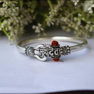 May include: Silver-toned bracelet with a central decorative element featuring the word "Mahadev" in a stylized script. The bracelet has a textured band and is accented with a red bead. The background includes white flowers.