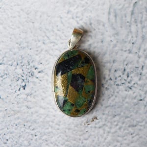Peut inclure: Pendentif ovale avec une bélière argentée. Le pendentif présente un motif en mosaïque avec des sections dorées, vertes et noires. La pierre est sertie dans une lunette en argent. Le design général est géométrique et accrocheur.