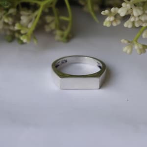 May include: A silver ring with a rectangular face. The ring is made of a shiny, polished metal and has a simple, modern design. The ring is set against a white background with some greenery in the background.