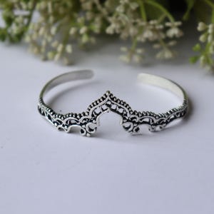 Peut inclure: Un bracelet manchette en métal argenté au design ouvert, orné de motifs filigranes symétriques complexes et d'un élément central pointu en forme de couronne. Le métal présente une finition oxydée sombre dans les creux pour souligner les détails.