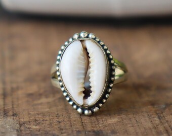 Könnte beinhalten: Ein Silberring mit einer weißen Kaurischnecke. Die Schnecke ist in einer silbernen Lünette mit einem Perlenrand gefasst. Der Ring liegt auf einer Holzoberfläche. Schmuck.