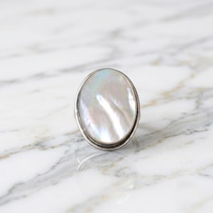 Peut inclure: Une bague ovale dotée d'un cabochon en nacre irisée lisse, serti dans une monture en métal argenté poli. La pierre précieuse présente des teintes blanches douces et nacrées avec de subtils reflets lumineux sur sa surface.