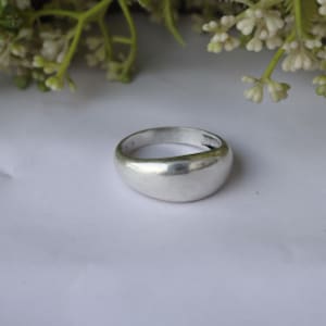 May include: A silver ring with a smooth, rounded design. The ring is made of a shiny, reflective metal and sits on a white surface. The ring's simple, yet elegant design makes it a versatile piece of jewellery.