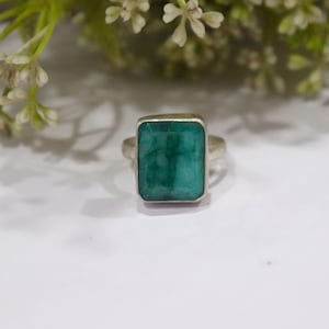 May include: A silver ring with a large, rectangular emerald-cut green gemstone. The ring has a simple design with a polished silver band and a bezel setting. The background is a soft white surface. The emerald is a vibrant shade of green.