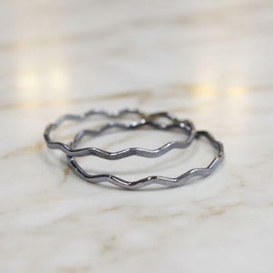 May include: Two thin, dark gray metal bangles with a wavy, undulating design. Each bracelet features a textured surface along the entire length of the band, creating a consistent, repeating pattern that follows the curves of the metal wire.