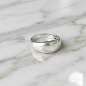 May include: A polished silver-toned ring with a smooth, rounded, and thick band. The metal surface is reflective and features a continuous, seamless design that curves gently around the finger opening, creating a solid and uniform appearance.