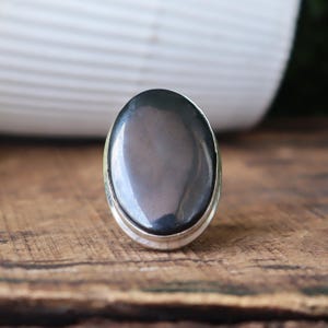 May include: A silver ring featuring a large, oval-shaped, polished black stone. The stone has a smooth, reflective surface. The ring is set on a wooden surface, with a blurred white background.
