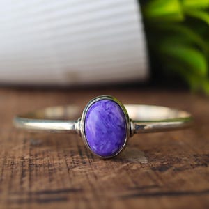 Pode incluir: Uma pulseira de prata com uma pedra preciosa oval roxa marmorizada. A pedra preciosa está engastada em uma moldura de prata. A pulseira é exibida em uma superfície de madeira.