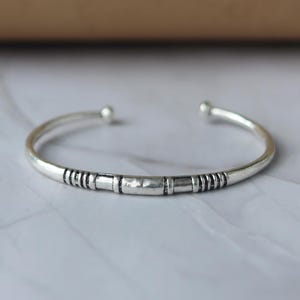 May include: A silver cuff bracelet with a central rectangular design and striped detailing. The open-ended bracelet has rounded tips. It is displayed on a white and grey surface.