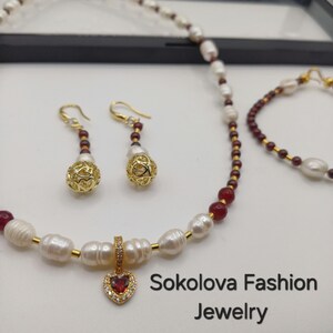 May include: A jewelry set featuring a necklace, earrings, and bracelet. The necklace and bracelet have white pearls and red beads. The earrings have gold filigree balls. The necklace has a heart-shaped pendant with a red stone. The text "Sokolova Fashion Jewelry" is visible.