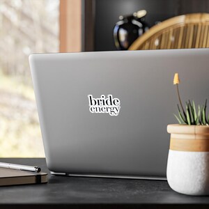 Könnte beinhalten: Ein grauer Laptop mit einem weißen Aufkleber mit der Aufschrift "bride energy" in schwarzer, moderner Schrift. Der Laptop steht auf einer schwarzen Oberfläche, rechts daneben eine kleine Topfpflanze.