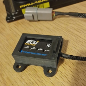 May include: Black ECU Master CAN-BUS Bluetooth Mod with a black cable and a grey connector. The device is on a wooden surface. Another black device with the text "PMU-16AS" is in the background.