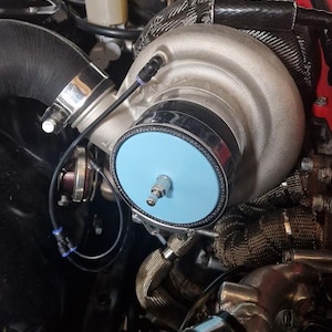 May include: A detailed view of a car engine featuring a silver turbocharger with a light blue circular element. The engine is filled with pipes, wires, and other components. The engine bay is dark with some red details.