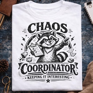 May include: White t-shirt with a black and white graphic of a raccoon holding a clipboard and pointing. The text on the shirt reads "CHAOS COORDINATOR - KEEPING IT INTERESTING."