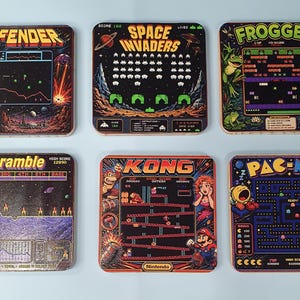 May include: A set of six square drink coasters featuring colourful graphic prints of classic arcade game screens including Defender, Space Invaders, Frogger, Scramble, Kong, and Pac-Man, each displaying its unique title, gameplay, and score.