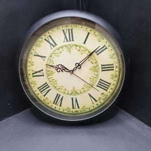May include: A round wall clock with a black frame and a cream-coloured face. The clock features black Roman numerals and ornate black metal hands. A decorative, light green filigree pattern encircles the centre of the clock face, providing a detailed contrast against the neutral background.