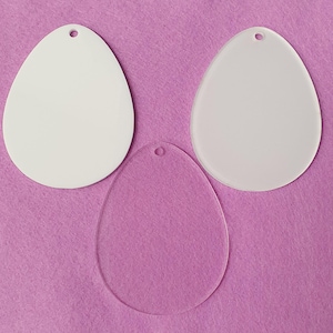 May include: Three egg-shaped blank ornaments. Two are opaque white, and one is transparent. Each has a small hole at the top for hanging. The ornaments are displayed on a pink felt surface.