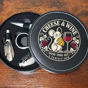 May include: A round black wine tool set case with white stitching. The lid features a graphic of a mouse holding cheese next to a wine glass, with the text CHEESE & WINE WINE TOOL SET A PERFECT PAIR. Inside are a bottle stopper, pourer, and corkscrew.