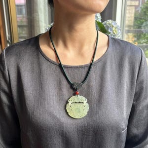 Puede incluir: Un colgante de jade tallado en color verde pálido suspendido de un cordón verde oscuro. El colgante circular presenta intrincados calados y está acentuado por una pequeña cuenta roja y un detalle de nudo negro decorativo en la parte superior.
