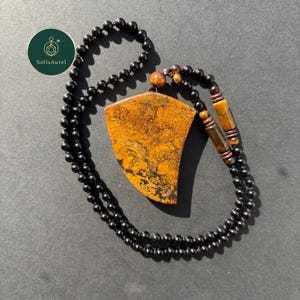 以下が含まれることがあります： オレンジと黒のまだら模様の大きな盾型のペンダントが付いたネックレス。ペンダントは小さな黒いビーズの紐から吊り下げられています。ペンダントの近くには、追加のビーズと木製のアクセントも見えます。