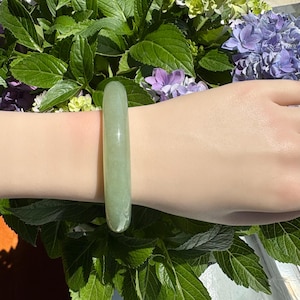 May include: A smooth, solid, light green jade bangle bracelet with a polished finish. The circular band features a consistent thickness and a translucent appearance, showcasing the natural pale green coloration of the stone material.