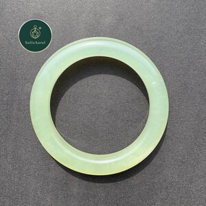 May include: A pale green jade bangle bracelet. The circular bracelet is smooth and polished, with a translucent appearance. The SolisAurel logo is in the upper left corner. The background is a dark gray.
