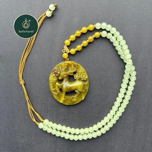 May include: A jade pendant necklace featuring a carved deer design within a floral border. The pendant is olive green, suspended from a strand of yellow and light green beads. The necklace has a gold-colored cord.