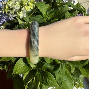 Könnte beinhalten: Ein glatter, polierter Armreif mit einem Marmormuster aus Wirbeln in dunklem Anthrazitgrau und cremigem Off-White. Der Armreif hat ein abgerundetes, zylindrisches Profil und eine glänzende Oberfläche, die das Licht auf der Außenseite reflektiert.