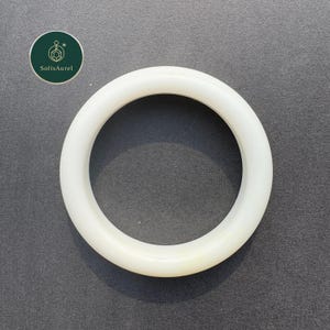 May include: A white, circular bangle bracelet. The smooth, solid bracelet is made of a light-colored material. The bracelet is set against a gray background. The brand logo is in the upper left corner.